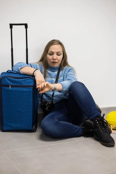 A woman at the airport sits on the floor with her Luggage and looks at her watch. The concept of a flight delay.