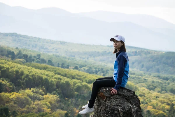 A girl traveler sits alone on a mountaintop against a background of forest and clouds.A woman on top of a mountain looks at the camera. The concept of tourism, travel and healthy lifestyle