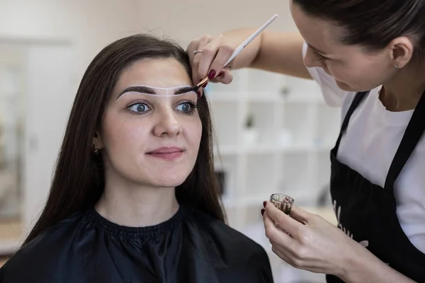 The master draws auxiliary lines between the eyebrows with a white pencil for permanent makeup. Creating an eyebrow sketch. Correction and coloring of eyebrows.