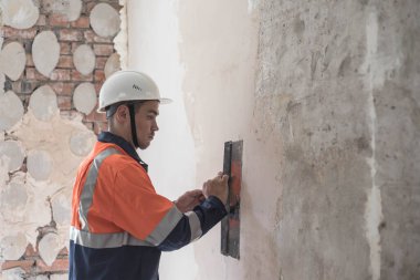 Kasklı ve iş üniformalı bir adam duvarları spatula ve macunla hizalıyor. Boşluğu kopyala