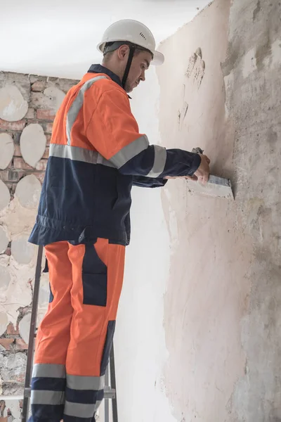 A construction worker in a white helmet and a work uniform is standing on a stepladder plastering the walls. A man is making repairs at home. The concept of repairing an apartment or house.