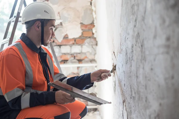 A man in a white helmet and work clothes works with a spatula, plastering the walls. The concept of apartment renovation. Free space for advertising.