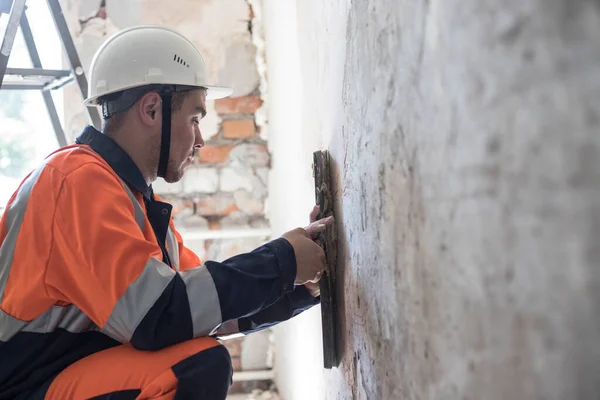 A man in a hard hat and work clothes is making repairs in an apartment room. Finishing works. Professional repair in the apartment. Plaster, painting, wall repair. Working with a spatula. Copy space.