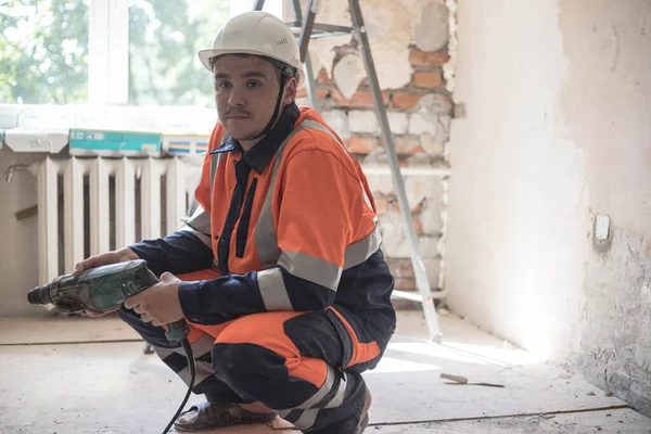 A man in a white helmet with a puncher in his hands makes repairs at home. Hammer drill.