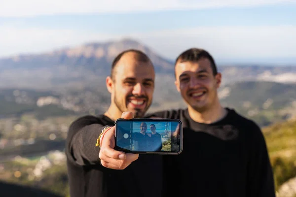Two friends taking a self-portrait with a mobile phone, on a sunny day with few clouds, on a mountain between villages, two dark-haired Caucasian boys in dark clothes.
