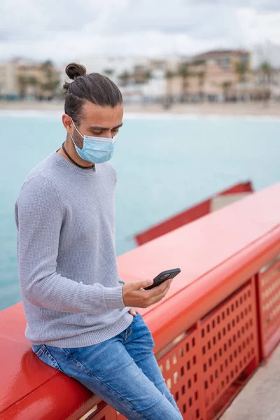 Kırmızı korkuluklara dayanan cerrahi maskeli bir cep telefonu kullanan bir adam. Bulutlu bir günde, gezinti güvertesinde. Onun bir eli cebinde. Kot pantolon giyiyor, gri bir kazak. alicante, javea