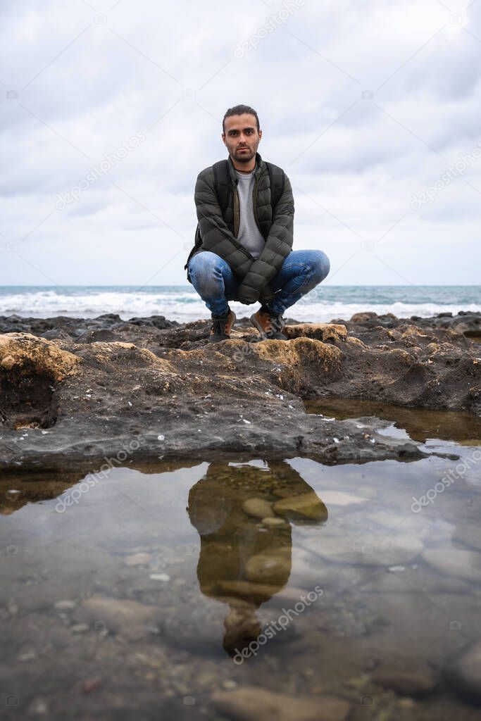 Squatting man reflected in a puddle in the sea. On a cloudy day. He is wearing a green jacket, grey sweater and jeans. Caucasian, with ponytail and rucksack on his back.