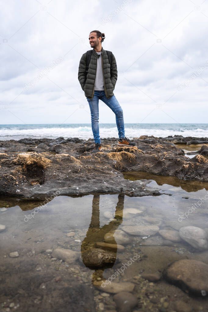 Happy man reflected in a puddle in the sea. On a cloudy day. He is wearing a green jacket, grey sweater and jeans. Caucasian, with ponytail and rucksack on his back.