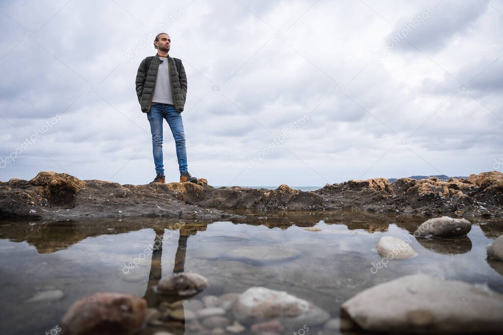 Man reflected in a puddle in the sea. On a cloudy day. He is wearing a green jacket, grey sweater and jeans. Caucasian, with ponytail and rucksack on his back.