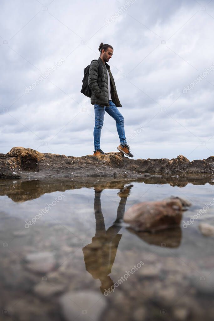 Man reflected in a puddle in the sea. On a cloudy day. He is wearing a green jacket, grey sweater and jeans. Caucasian, with ponytail and rucksack on his back.