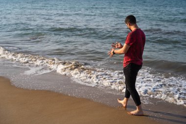 Avrupalı bir adam kumsalda ellerini kumdan sallıyor. Güneşli bir günde plajın kıyısında yürüyor. Kırmızı tişört, siyah pantolon, saat ve maske giyiyor. Akdeniz 'de, Denia.