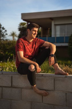 Duvarda oturan adam gülümsüyor. O yalınayak ve bir saat takıyor. Kırmızı kısa kollu tişört ve siyah pantolon giyiyor..