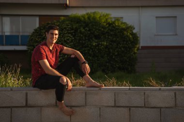 Duvarda oturan adam gülümsüyor. O yalınayak ve bir saat takıyor. Kırmızı kısa kollu tişört ve siyah pantolon giyiyor..