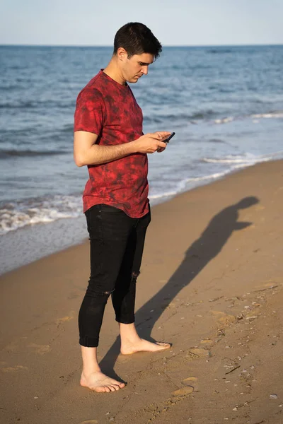 Deniz kıyısındaki adam. O, iki eliyle cep telefonunu kullanıyor. Güneşli bir günde. Kafkasyalı ve koyu renk saçlı. Kırmızı kısa kollu tişört ve siyah pantolon giyiyordu. O yalın ayak..