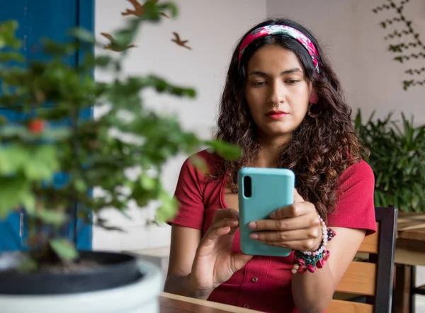Kırmızı gömlekli ve başörtülü genç Latin kadın kafede oturuyor ve telefon kullanıyor.