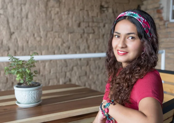 Restoranda oturan ve gülümseyen kırmızı tişörtlü ve başörtülü genç Latin kadın.