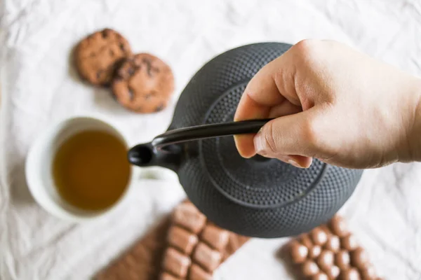 Cup of tea, a hand holding an iron teapot and cookies on the white cloth background, morning food and drink, sweet pastry dessert with chocolate and nuts