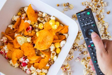 Beyaz arka planda kızarmış patlamış mısır ve cips, el ele tutuşan televizyon kumandası, düşük açılı görüş.