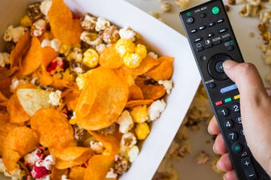 Beyaz arka planda kızarmış patlamış mısır ve cips, el ele tutuşan televizyon kumandası, düşük açılı görüş.