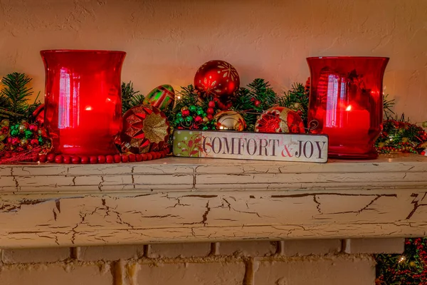 A plaque that says comfort and joy sits on a fireplace mantel surrounded by two hurricane candles and Christmas ornaments.