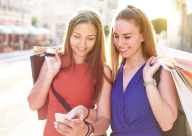 Genç mutlu milenyum arkadaşları alışveriş çantasıyla sokaklarda yürüyor ve sosyal medya platformlarının güncellemelerini kontrol ediyor - iki öğrenci bir web dükkanından kıyafet sipariş ediyor - arka planda ışıklandırmalı alışveriş konsepti