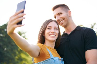 Genç mutlu milenyum çifti gülümsüyor ve selfie çekiyor - Z jenerasyonu turistler birlikte eğleniyor - akıllı telefon ve arkadaşlarıyla teknoloji kavramı