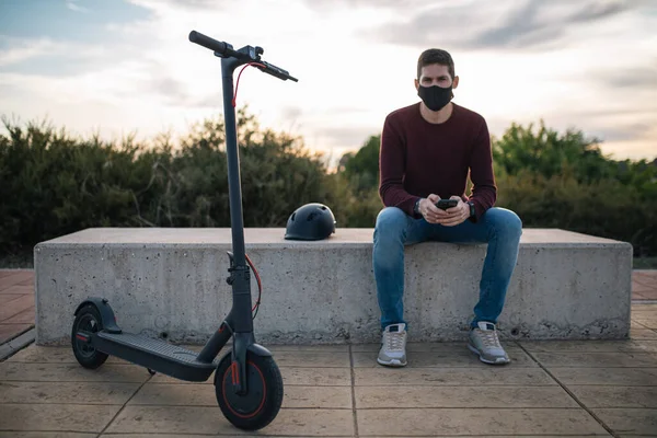 Young man on an electric skateboard using a mobile phone sitting on a bench with his helmet. Alternative transport.