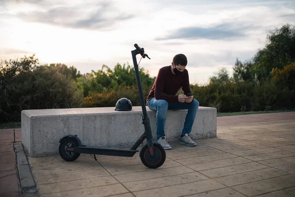 Young man on an electric skateboard using a mobile phone sitting on a bench with his helmet. Alternative transport.