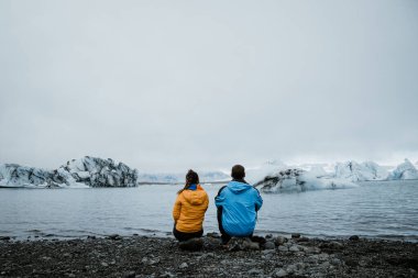 Tanımlanamayan çift sırtüstü oturuyor. İzlanda 'daki bir buzula bakıyorum. Sarı ve mavi paltolar. Metin için seyahat ve aşk alanı kavramı.