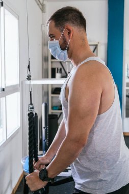 Spor salonunda maskeli bir adam ipten trisepsler yapıyor. Covid19. Coronavirüs