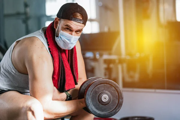 Man with mask doing biceps exercises with dumbbells in the gym. sitting on a bench. Covid19, coronavirus. Health and wellness concept