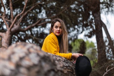 Güzel bir kadın ağaca yaslanmış sarı bir palto ve siyah bir şapka giyiyor.