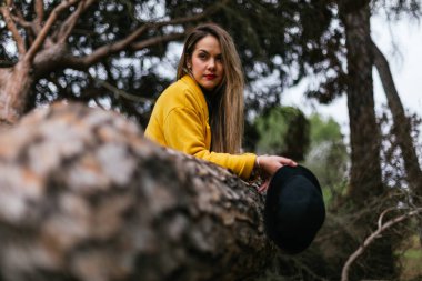 Güzel bir kadın ağaca yaslanmış sarı bir palto ve siyah bir şapka giyiyor.