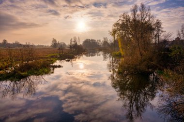 Nehirdeki ağaç ve bulut yansımalarının arka planda çekilmiş bir fotoğrafı. Arka planda güzel bir günbatımı gökyüzü. Fotoğraf İsveç 'in İskandinav ilçesinden.