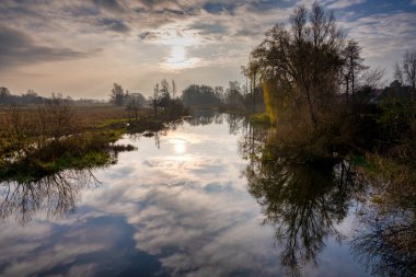 Nehirdeki ağaç ve bulut yansımalarının arka planda çekilmiş bir fotoğrafı. Arka planda güzel bir günbatımı gökyüzü. Fotoğraf İsveç 'in İskandinav ilçesinden.