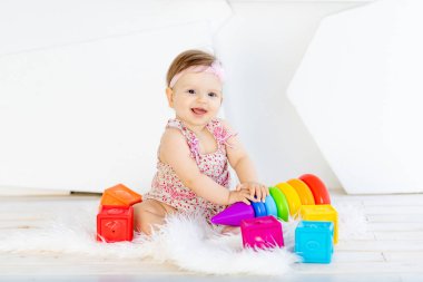 Kırmızı elbiseli, beyaz bir odada parlak oyuncaklarla oturan mutlu küçük kız.