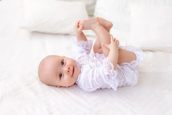 一个6个月大的小女孩穿着漂亮的白色紧身衣躺在轻便的床上 对着摄像机笑着 宝宝仰卧在床上 两腿紧抱着 — 图库照片