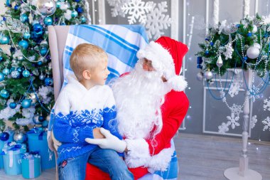 Noel Baba, mavi bir Noel ağacının yanında kucağında bir bebek tutuyor. Mutlu bir çocuk Noel Baba 'yla konuşuyor.