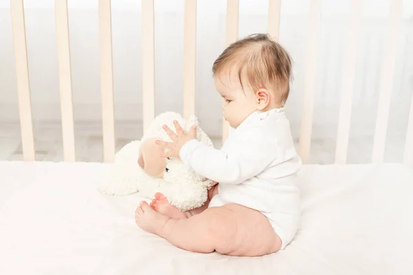 6个月大的婴儿穿着白色紧身衣和泰迪熊在婴儿床里玩耍 — 图库照片