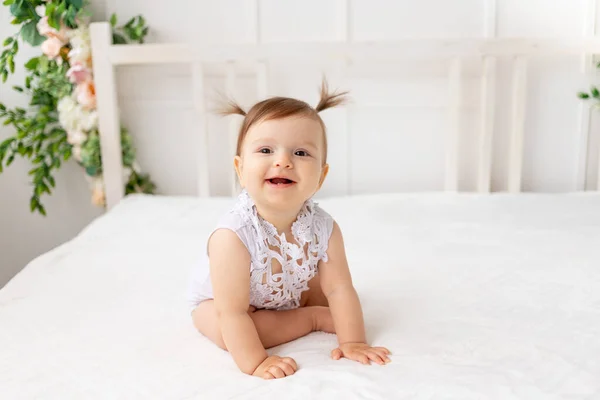 6个月大的小女孩坐在一个明亮美丽的房间里 白色的床 身穿花边紧身衣 看着相机 面带微笑 — 图库照片