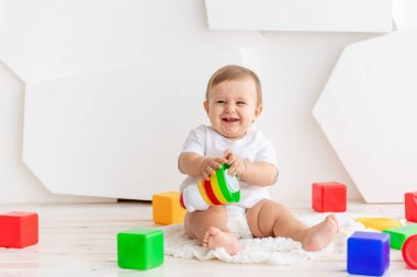 6 aylık beyaz tişörtlü ve bez bezli mutlu bir bebek. Parlak renkli küplerle parlak bir odada, Mat 'in üzerinde oynuyorlar.
