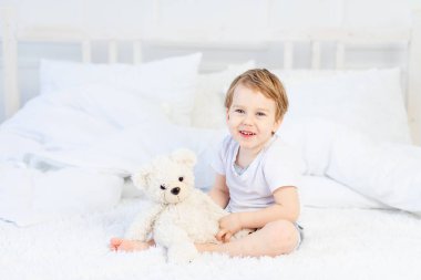 Evde yatağında oyuncak ayıyla oynayan ve gülen bir çocuk.