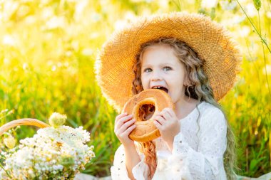 Sarı bir tarlada hasır bir şapkada oturan küçük güzel bir kız yanında bir şişe süt ve simit ile yabani çiçeklerle tarlada piknik yapıyor.
