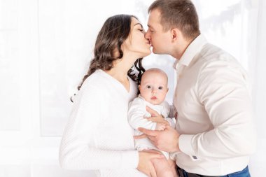 Anne ve baba yeni doğmuş bir bebeği pencerede tutarak öpüşüyorlar. Mutlu sevgi dolu aile kavramı, yaşam tarzı.