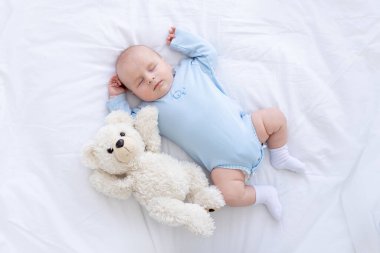 Bebek çocuk, mavi pijamalı yumuşak bir oyuncak ayıyla sırt üstü yatar ve ellerini kaldırıp sağlıklı bir şekilde uyur.