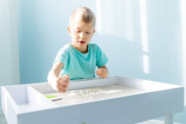 Çocuk evde kumla oynuyor masada, kumları çiziyor.