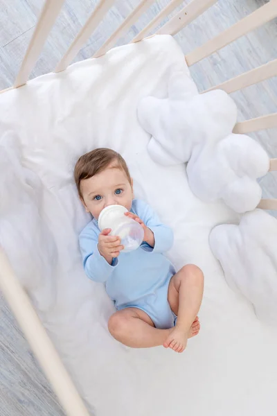ベビーベッドでミルクボトルから青いボディスーツで食事寝室でかわいい赤ちゃんベビーフードコンセプト — ストック写真