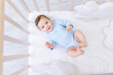 Mavi elbiseli beşikteki mutlu erkek bebek, yatak odasında sevimli, neşeli küçük bebek
