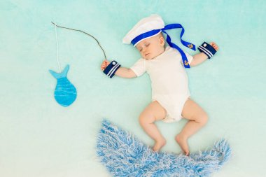 Mavi arka planda bir erkek bebek denizci kıyafetiyle uyur ve rüyasında dalgalarda yüzer ve balık yakalar.