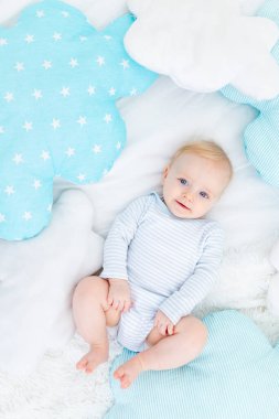 Bebek altı aylık, yumuşak mavi yastıklı bir yatakta uzanmış, gülümseyen sarışın bir bebek.